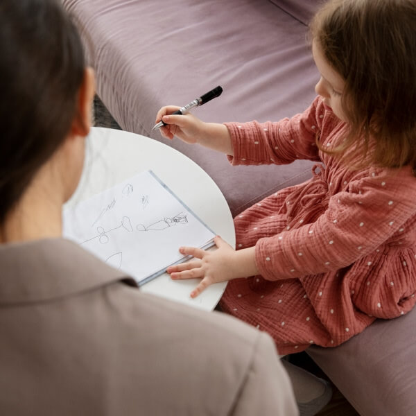 Une vue en plongée d'une jeune fille en robe rose assise sur un canapé violet et dessinant avec un stylo sur du papier, sous l'observation d'une spécialiste.